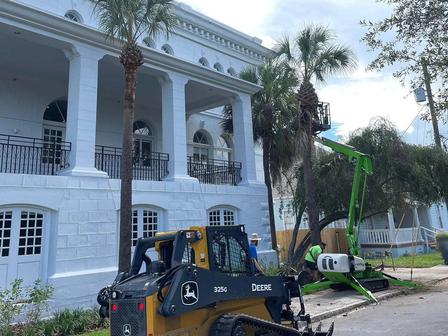Commercial tree service in Tampa, FL: a tracked lift and a compact track loader working among palms beside a large white building with columns