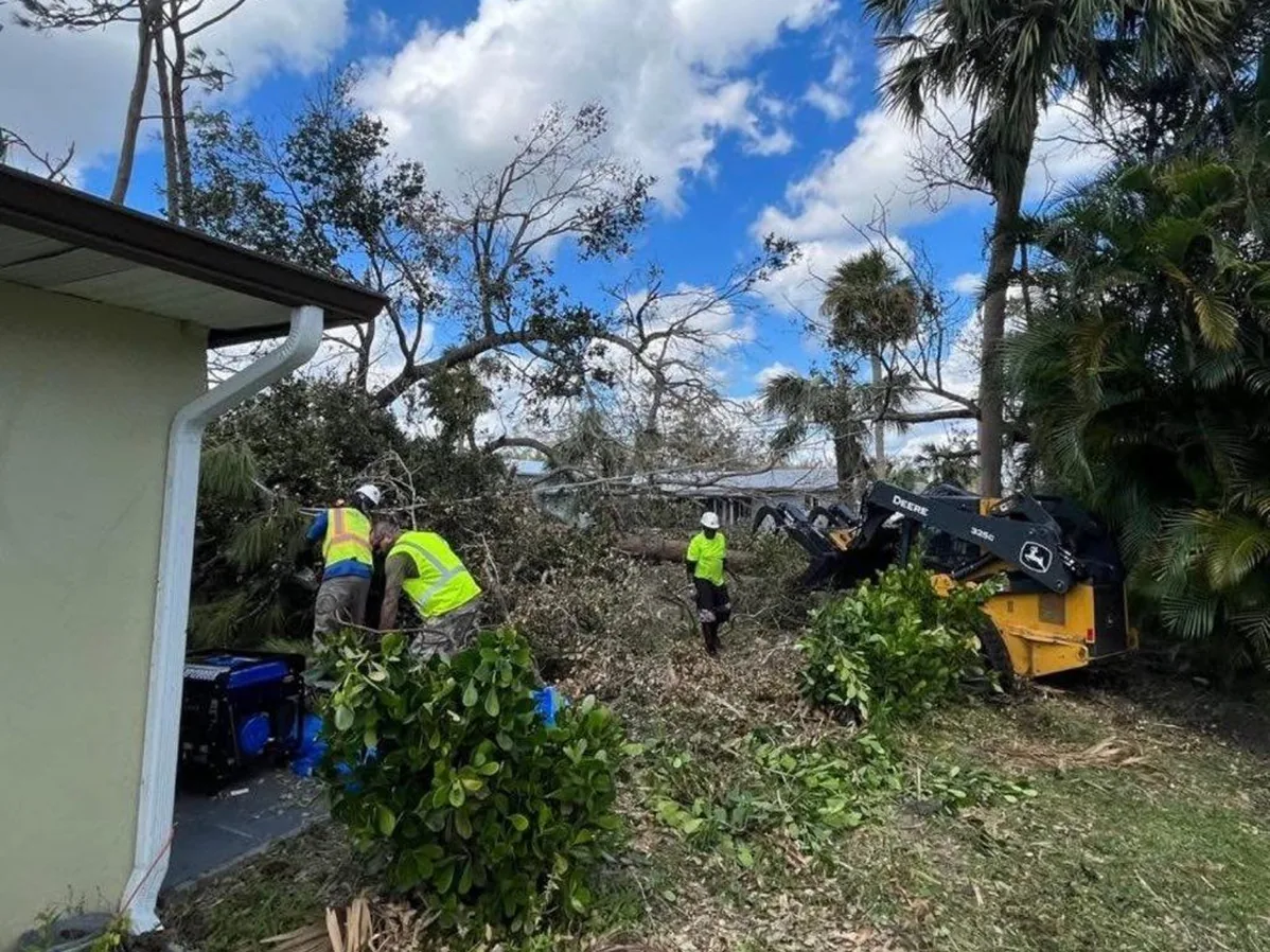 Storm cleanup: a crew in high-vis cutting and dragging fallen trees beside a house, with a compact loader and a grapple