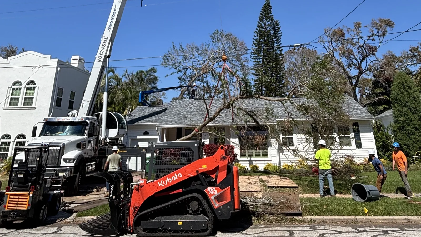 Tree removal on a residential street: a truck-mounted crane rigged to a tree section above a house, with a compact track loader, a grapple and a crew clearing brush