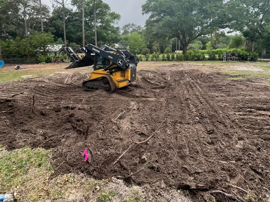 Site preparation: a compact track loader rough-leveling a cleared and grubbed lot, with oaks left standing at the edge