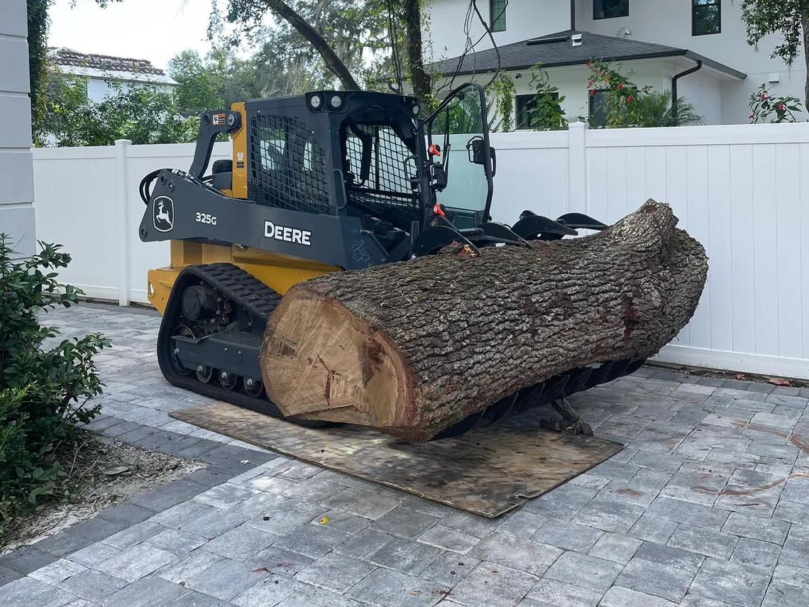 Tree removal cleanup: a compact track loader with a grapple carrying a large trunk section across a paver driveway