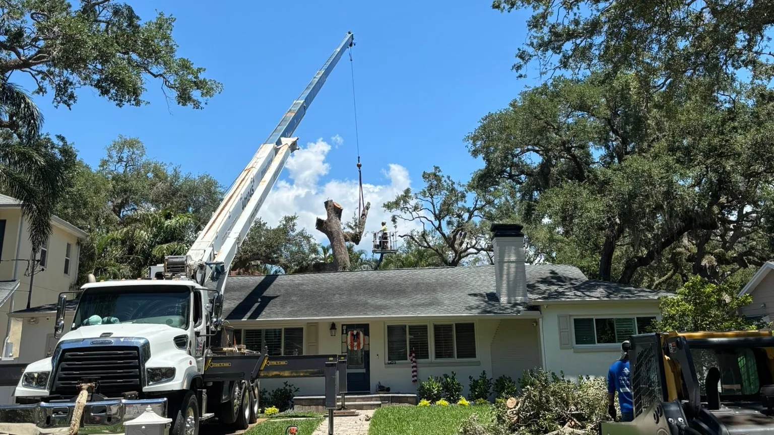 A truck-mounted crane set up in front of a house for a tree removal, with a compact track loader beside it